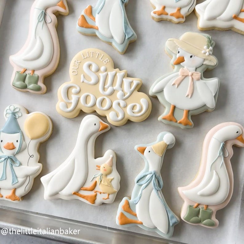 May include: A collection of decorated sugar cookies in the shape of geese. Some cookies feature geese wearing hats, bows, and boots. One cookie has the text "Our Little Silly Goose". The cookies are white, yellow, orange, pink, and blue.