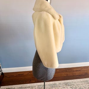 May include: A cream-colored faux fur jacket with a wide collar and cropped length. The jacket is displayed on a gray dress form with a metal stand. The background has a gradient of blue and white.