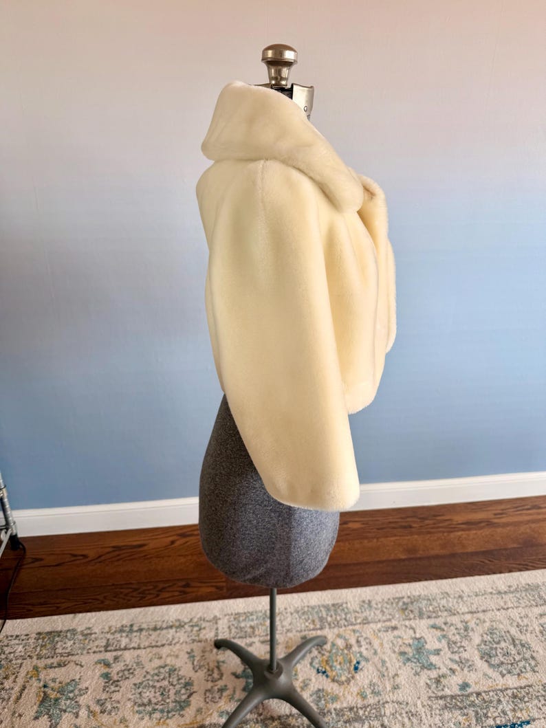 May include: A cream-colored faux fur jacket with a wide collar and cropped length. The jacket is displayed on a gray dress form with a metal stand. The background has a gradient of blue and white.