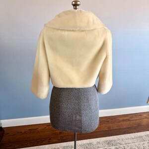 May include: A cream-colored, cropped faux fur jacket with a wide collar and three-quarter sleeves. The jacket is displayed on a dress form with a gray skirt. The background has a gradient of blue and pink.