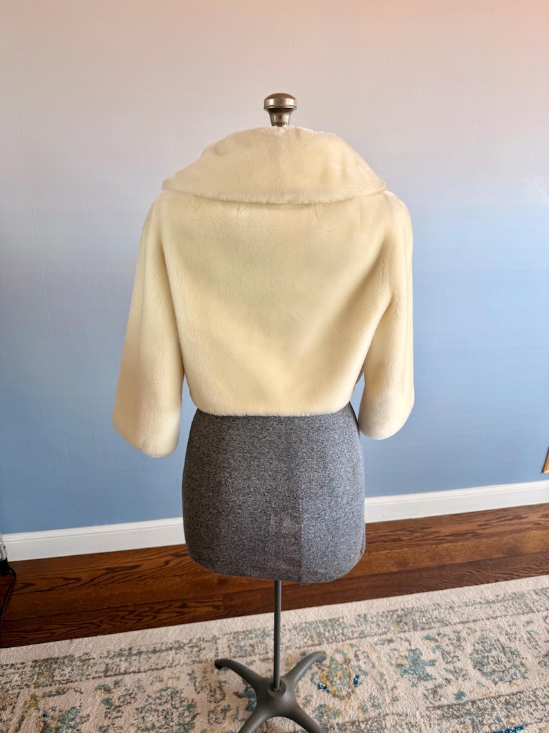 May include: A cream-colored, cropped faux fur jacket with a wide collar and three-quarter sleeves. The jacket is displayed on a dress form with a gray skirt. The background has a gradient of blue and pink.