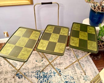 May include: A set of three rectangular folding snack tables with gold-tone metal legs and frames. Each tabletop features a checkered pattern of dark green and light olive green squares with a textured finish. A matching gold-tone storage rack is included.