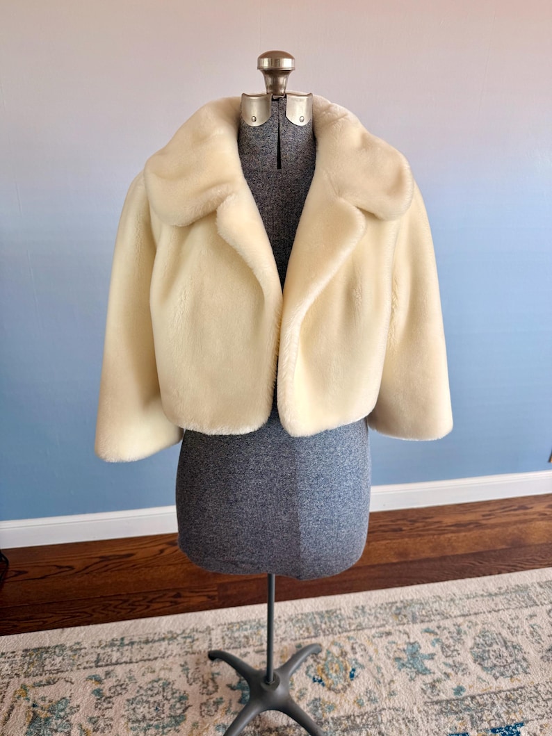 May include: Cream-colored faux fur cropped jacket with a wide collar and three-quarter sleeves. The jacket is displayed on a gray dress form against a blue and white background. The jacket's soft texture and classic design are highlighted.