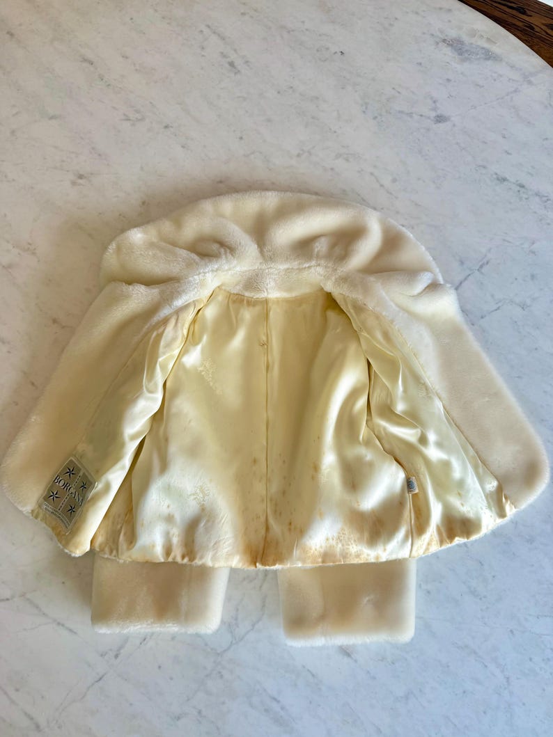 May include: Cream-colored faux fur coat with a hood and satin lining. The coat has a classic design with a soft, plush exterior and a smooth, light yellow interior. The coat has a label that reads "BORG".