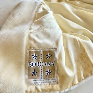 May include: Cream-colored faux fur coat with a satin lining. The interior lining is a light cream color with a subtle pattern. A label reads "BORGANA DELUXE" with text below that says "CLEAN THIS FABRIC BY FUR COAT METHOD DO NOT BRUSH, CARD, PRESS OR STEAM."