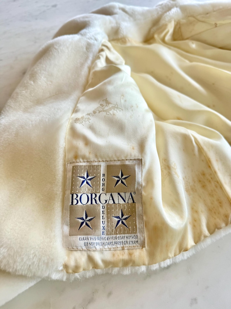 May include: Cream-colored faux fur coat with a satin lining. The interior lining is a light cream color with a subtle pattern. A label reads "BORGANA DELUXE" with text below that says "CLEAN THIS FABRIC BY FUR COAT METHOD DO NOT BRUSH, CARD, PRESS OR STEAM."