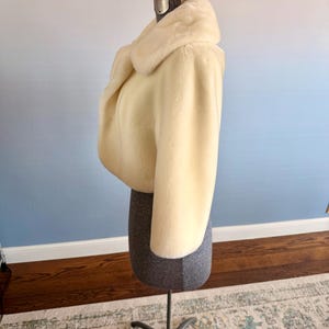 May include: Cream-colored faux fur bolero jacket with a wide collar. The cropped jacket is displayed on a gray dress form with a metal stand. The background is a gradient of blue and white.
