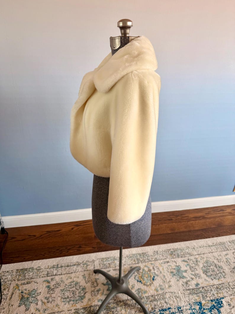 May include: Cream-colored faux fur bolero jacket with a wide collar. The cropped jacket is displayed on a gray dress form with a metal stand. The background is a gradient of blue and white.