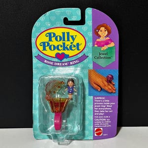 May include: A Polly Pocket Rose Dream Ring Jewel Collection toy set. The packaging is teal, purple, and pink, with a clear plastic window showing the ring and a small doll. The ring is pink and has a small, enclosed playset.