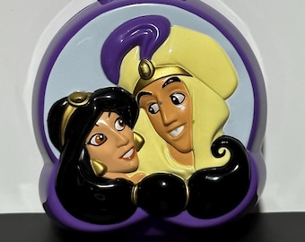 Könnte beinhalten: Ein violetter Plastik-Kompakt mit einem Bild von Aladdin und Jasmin. Der Kompakt hat einen goldenen Rand und einen weißen Hintergrund.