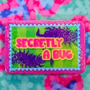 Può includere: Una spilla rettangolare con un bordo smerlato rosa glitterato e una cornice interna azzurra. Il centro presenta uno sfondo verde brillante con sagome di insetti viola scuro e il testo giallo "SECRETLY A BUG" in un carattere grassetto.