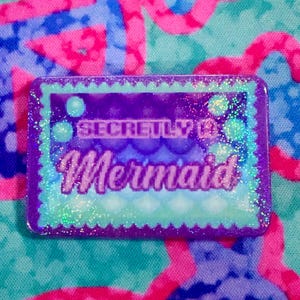 May include: A rectangular pin with a scalloped edge, featuring a purple and teal gradient background with a subtle scale pattern. The text reads "SECRETLY A Mermaid" in a mix of block and script fonts, all covered in a sparkling glitter finish.