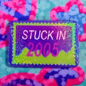 May include: A rectangular pin with a scalloped edge featuring a holographic finish. The design includes a purple background with a lime green accent at the bottom. The text STUCK IN 2005 is printed in white and purple across the center of the pin.