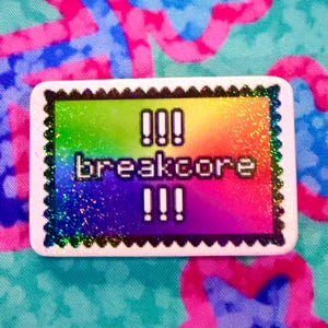 Puede incluir: Un pin rectangular con borde festoneado que presenta un fondo degradado de arcoíris y un acabado holográfico. El centro muestra el texto blanco pixelado "!!! breakcore !!!" en un estilo de fuente negrita y cuadrada, creando un efecto visual vibrante.