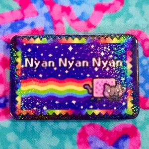 May include: A rectangular pin featuring a pixelated cat with a pink body trailing a rainbow across a dark blue starry background. The text Nyan Nyan Nyan is printed in white pixelated font. The border has a scalloped edge with a rainbow glitter finish.