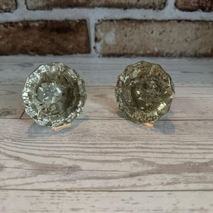 May include: Two clear, faceted glass cabinet knobs with a floral design. The knobs are round with a scalloped edge and a central flower pattern. They are displayed on a light wood surface.