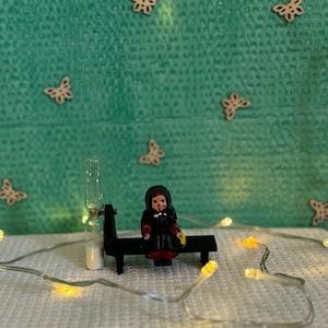 May include: A small doll wearing a black dress and a yellow scarf sits on a black bench. A clear glass bottle with a cork stopper sits next to the bench. The scene is lit by a string of white lights.