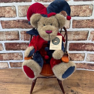 May include: A plush teddy bear wearing a red and navy blue jester hat and matching outfit. The bear has brown fur, a striped nose, and a tag that reads "Boyds Bear & Friends" and "Mr. Sparkle". It is seated on a small wooden chair.