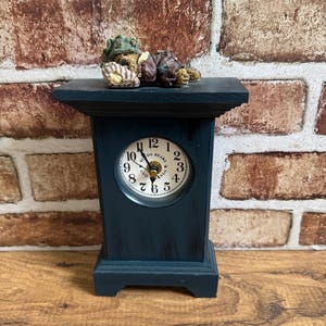 May include: A dark blue wooden mantel clock with a round white face featuring black numerals and the text "Boyds Bears and Friends". A small figurine of a bear wearing a green hat and brown clothing lies on top of the clock, resting on a pillow.