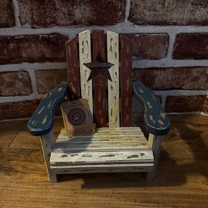 May include: A miniature wooden Adirondack chair featuring a distressed paint finish in red, white, and blue. The backrest displays a dark star cutout, and a brown cardboard tag with the text "GENUINE BOYDS" and a paw print logo is attached.