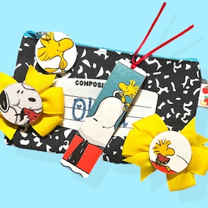 May include: A collection of Peanuts-themed accessories, including a black and white composition notebook-style zippered pouch, a bookmark, and several yellow bow hair clips. The bookmark and hair clips feature Snoopy and Woodstock characters.