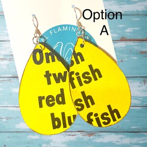 May include: Yellow teardrop earrings with the text "One fish, two fish, red fish, blue fish" in black. The earrings have silver-colored hooks and are hanging on a white card with a blue and white logo.