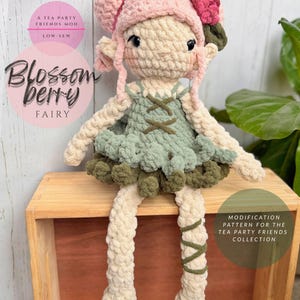 May include: A handmade crochet fairy doll with a pink cap, green dress, and cream-colored limbs. The doll has black eyes and is sitting on a wooden box. The text "Blossom Berry Fairy" is displayed on the image.