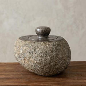May include: A round, stone-like ceramic container with a rough, textured grey and brown exterior. It features a smooth, dark grey circular lid with a small, rounded knob handle centred on top, creating a contrast between the coarse base and lid.