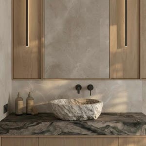 Puede incluir: Un mueble de baño moderno con un lavabo de piedra. La encimera es oscura con un patrón de mármol y los armarios son de madera clara. Se ven dos grifos negros y dos apliques de pared a juego.