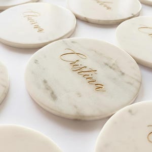 Puede incluir: Posavasos redondos de mármol blanco con letras doradas. Los posavasos tienen un acabado pulido y están grabados con nombres en una fuente cursiva. El mármol tiene vetas grises. Cada posavasos mide aproximadamente 10 cm de diámetro.