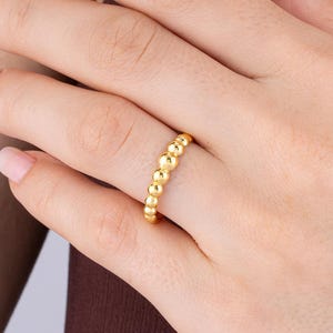 Puede incluir: Un anillo de oro que presenta una banda continua de pequeñas cuentas esféricas pulidas. El metal tiene un acabado brillante y reflectante, y el diseño consiste en una serie de elementos redondeados uniformes unidos para formar un anillo.