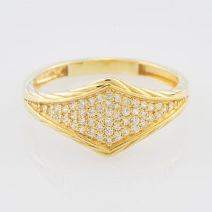 Puede incluir: Un anillo de oro con un centro en forma de diamante lleno de pequeñas piedras preciosas claras. El anillo tiene una banda texturizada y un acabado pulido, que refleja la luz. El anillo está sobre un fondo blanco.