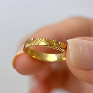 May include: A gold band ring featuring a repeating engraved pattern of sunbursts. The metal has a polished finish, and the design consists of multiple semi-circular sun motifs with radiating lines etched around the entire circumference of the ring.