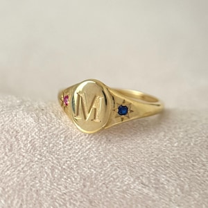 以下が含まれることがあります： 文字「M」が刻印された楕円形のフェイスを持つゴールドのシグネットリング。リングの肩部分には、中央の文字の両側にピンクとブルーの小さな星型にセットされた宝石があしらわれています。