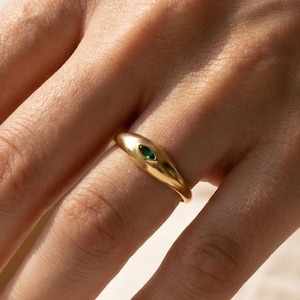 Puede incluir: Un anillo de oro con un diseño liso y curvado. El anillo presenta una pequeña gema verde de forma ovalada engastada en la banda. El anillo se muestra en el dedo de una persona.