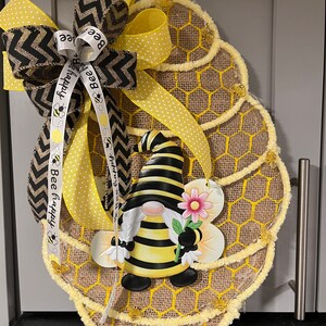 May include: Bee-themed burlap wreath featuring a gnome dressed as a bee, holding a flower. The wreath is decorated with yellow and black chevron ribbon, yellow polka dot ribbon, and a ribbon with the text 'Bee Happy'.  The wreath's base is made of burlap with a honeycomb pattern and yellow accents. Small bee embellishments are also present.