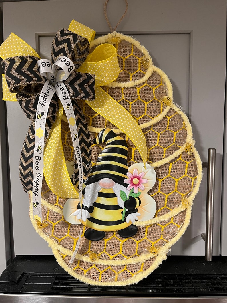 May include: Bee-themed burlap wreath featuring a gnome dressed as a bee, holding a flower. The wreath is decorated with yellow and black chevron ribbon, yellow polka dot ribbon, and a ribbon with the text 'Bee Happy'.  The wreath's base is made of burlap with a honeycomb pattern and yellow accents. Small bee embellishments are also present.