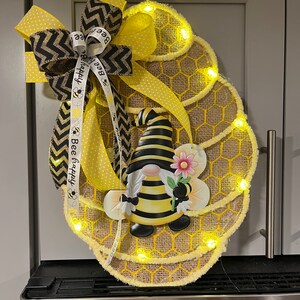May include: A bee-themed spring wreath features a burlap honeycomb design accented with yellow polka dot ribbon and black and yellow chevron ribbon.  A charming bumblebee gnome is centered, holding a pink flower.  The wreath is illuminated by small, warm-toned LED lights embedded along the edges of the honeycomb.  Ribbons display the text 'Bee happy'.