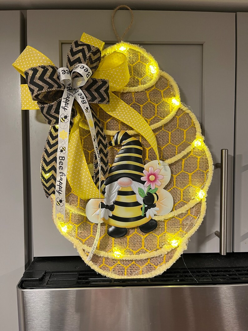 May include: A bee-themed spring wreath features a burlap honeycomb design accented with yellow polka dot ribbon and black and yellow chevron ribbon.  A charming bumblebee gnome is centered, holding a pink flower.  The wreath is illuminated by small, warm-toned LED lights embedded along the edges of the honeycomb.  Ribbons display the text 'Bee happy'.