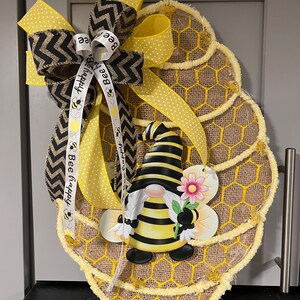 May include: Bee-themed burlap wreath featuring a gnome and honeycomb design.  Yellow and black color scheme with a chevron and polka dot ribbon. The ribbon includes the text 'Bee Happy'.  The wreath is accented with small yellow bee embellishments.