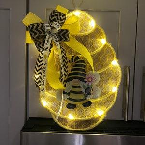 May include: A lighted burlap honeycomb-shaped wreath features a bumble bee gnome and a yellow and black chevron ribbon bow. The ribbon is adorned with the text 'Bee Happy'.  The wreath is decorated with small LED lights, creating a warm glow. The overall design evokes a cheerful, spring or summer theme.