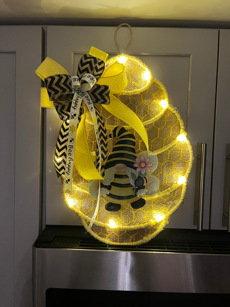 May include: A lighted burlap honeycomb-shaped wreath features a bumble bee gnome and a yellow and black chevron ribbon bow. The ribbon is adorned with the text 'Bee Happy'.  The wreath is decorated with small LED lights, creating a warm glow. The overall design evokes a cheerful, spring or summer theme.
