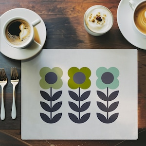 May include: A white placemat with a floral design featuring three stylized flowers in shades of green and blue, each with a dark grey centre and stem. The placemat is on a wooden table, with coffee cups and cutlery.