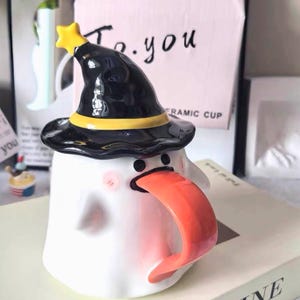 Puede incluir: Taza de cerámica blanca con forma de fantasma, con un sombrero de bruja negro con una banda y una estrella amarillas. La taza tiene un asa naranja y mejillas rosas. La taza está sobre un libro.