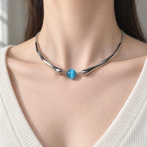 Puede incluir: Un collar de tono plateado que presenta una cuenta esférica central de color azul brillante con efecto ojo de gato, flanqueada por dos componentes metálicos lisos y alargados en forma de gota que se conectan a una fina cadena plateada.