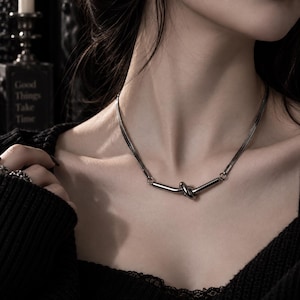 Puede incluir: Un collar de tono plateado con una cadena plana de espiga que se transforma en dos barras metálicas cilíndricas y lisas conectadas por un nudo central. El collar descansa sobre el cuello y muestra un acabado metálico pulido.