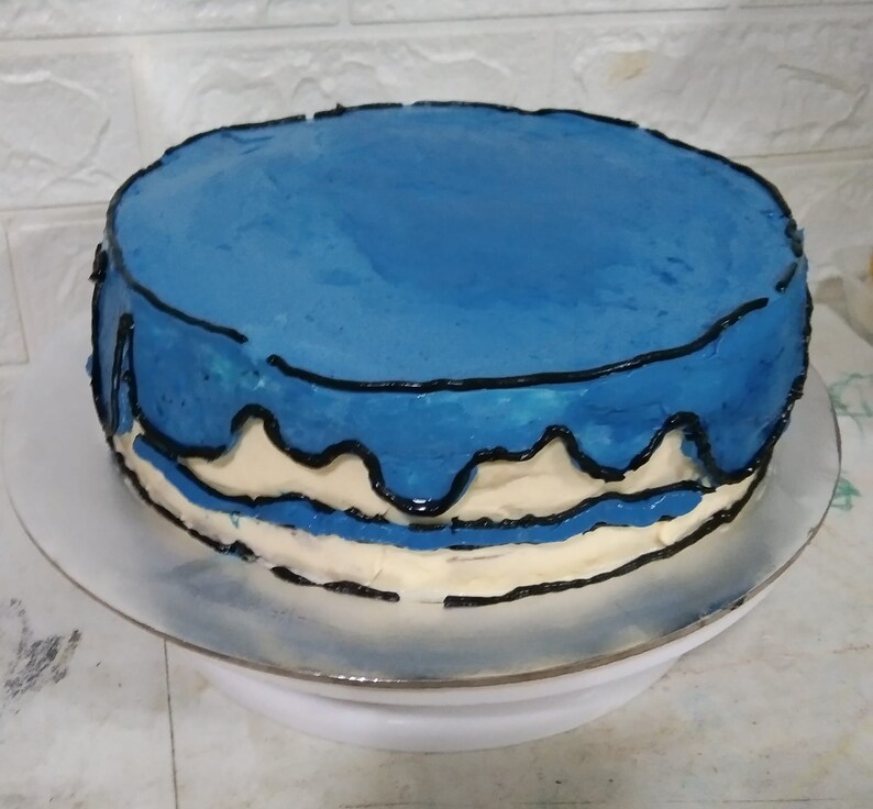Pode incluir: Um bolo glaciado azul e branco com glac&ecirc; preto pingando ao redor das bordas. O bolo est&aacute; em um suporte para bolo branco.