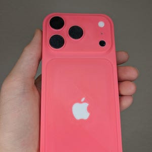 May include: A bright pink phone case with a white Apple logo on the back. The case has a rectangular shape with rounded corners and a raised section for the camera lenses. The camera section has three black circles and a white circle.