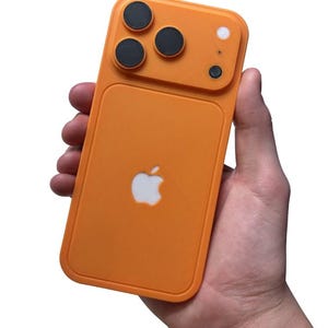 May include: Orange phone case with a white Apple logo. The case has a camera module with three black circles and a white circle. The case is held in a hand.