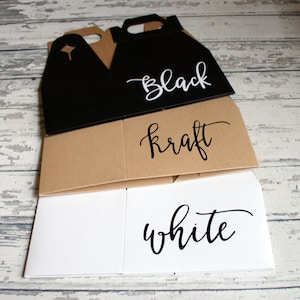 May include: Three empty beverage carrier boxes, one black, one brown kraft, and one white. The boxes are stacked on top of each other. The words "black", "kraft", and "white" are written on the boxes in black script.